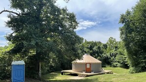 Uma yurt com porta de madeira e pequeno alpendre, rodeada por vegetação exuberante e árvores.
