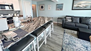 Eine moderne Küche mit Marmorarbeitsplatte, weißen Schränken und einer eingebauten Mikrowelle. Es gibt eine Essecke mit Barhocker und einem kleinen Tisch. Das Wohnzimmer bietet ein graues Sofa und einen blauen Teppich.