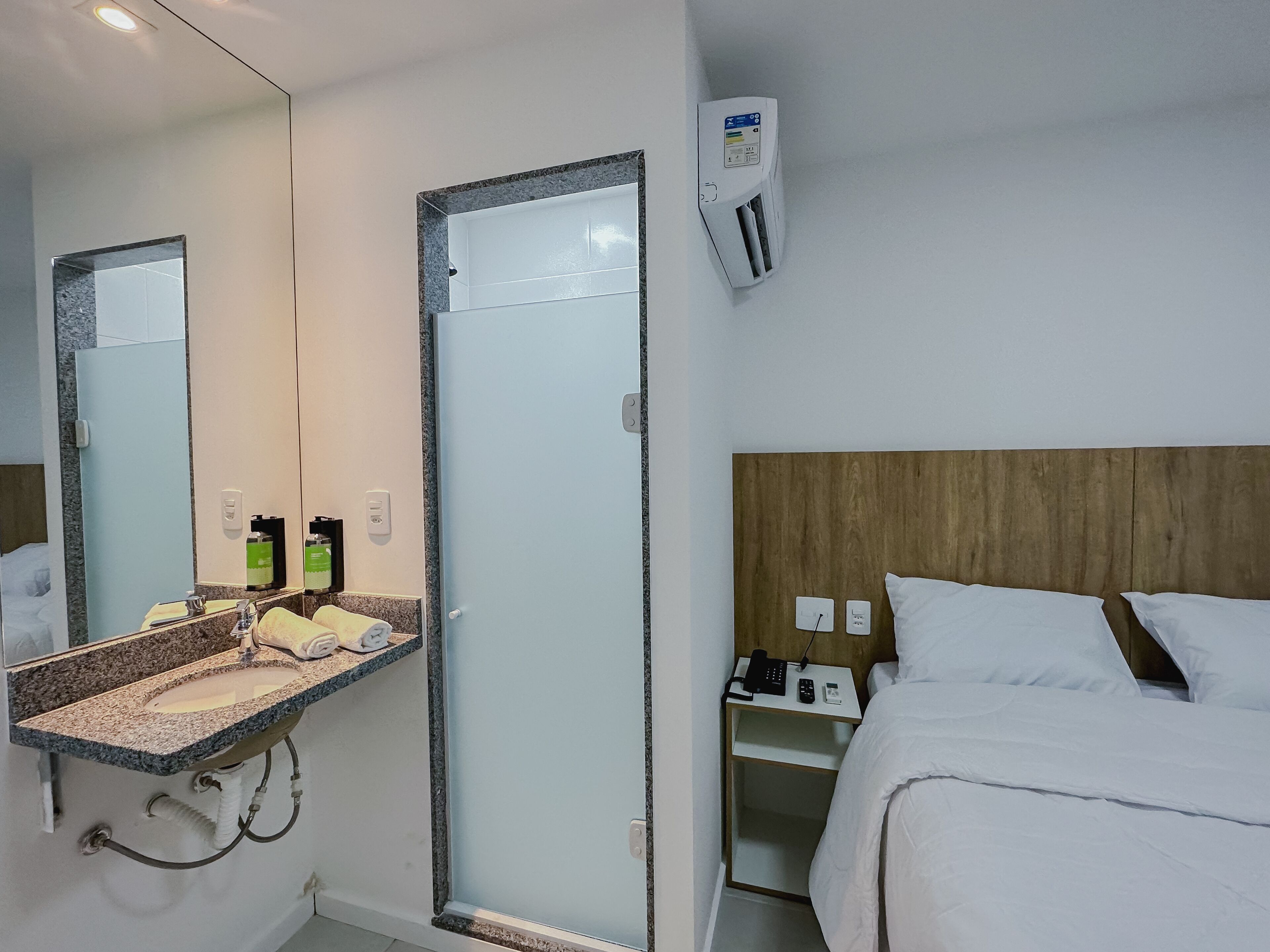 Habitación de hotel con un espejo grande, un lavamanos con artículos de aseo, una cama con ropa blanca y un cabecero de madera.