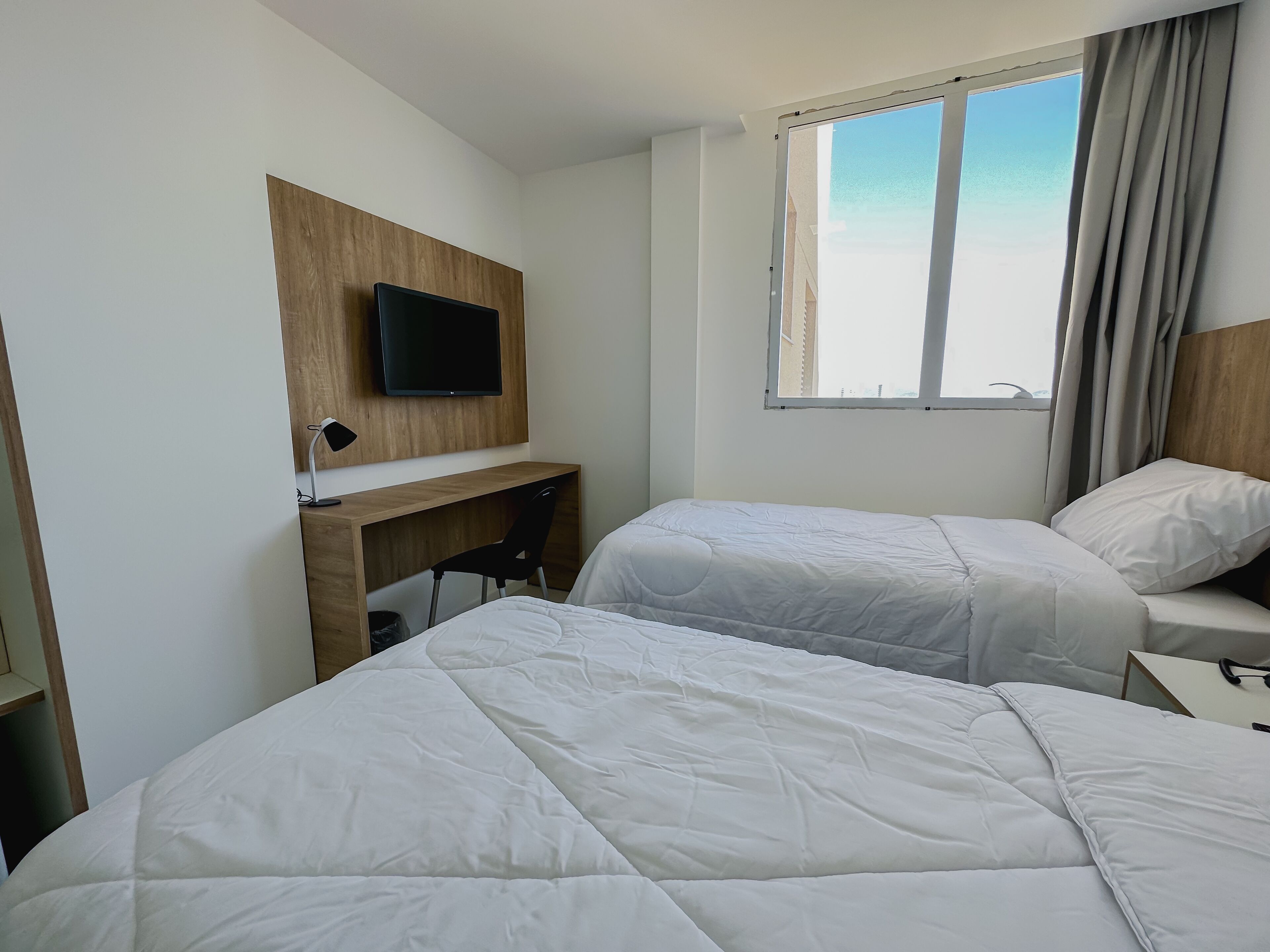 Quarto de hotel com duas camas, uma mesa, uma televisão e uma janela com cortinas.