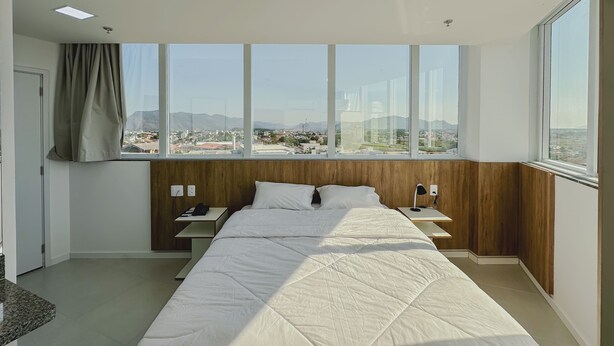 A modern hotel room with a large bed, a nightstand, a desk, and a view of a cityscape through large windows. - GO INN ARACRUZ (Aracruz)