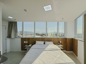 Habitación de hotel moderna con una cama grande, una mesita de noche, un escritorio y vistas a un paisaje urbano a través de amplias ventanas.