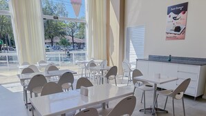 A clean, modern dining area with white tables and chairs, large windows with blinds, and a poster on the wall advertising a café. - GO INN ARACRUZ (Aracruz)