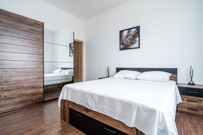Une chambre moderne avec une armoire en bois, un grand lit avec du linge de lit blanc, une table de chevet, une commode et un tableau abstrait encadré accroché au mur.