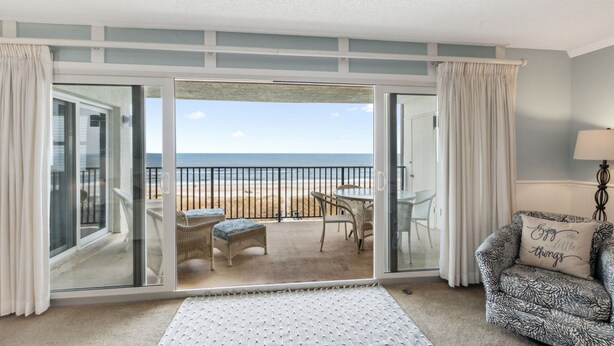 Une pièce dotée d’une porte-fenêtre coulissante donnant sur un balcon avec vue sur la plage, meublée de mobilier en osier et agrémentée d’une lampe.