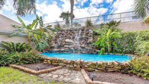 A backyard with a stone waterfall leading into a swimming pool, surrounded by lush greenery and palm trees. - Ocean Park 318 Harbor Point - Hosted By Moore AI Rentals (Fernandina Beach)