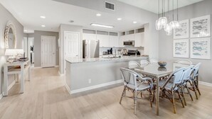 A modern kitchen with white cabinetry, a central island, and a dining area with wicker chairs. - Ocean Park 318 Harbor Point - Hosted By Moore AI Rentals (Fernandina Beach)