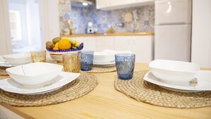 Una mesa de comedor dispuesta con platos blancos, tazones y vasos azules sobre individuales tejidos en una cocina con salpicadero de azulejos blancos y azules y un refrigerador blanco.
