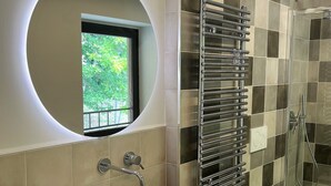 A modern bathroom with a round mirror, a vessel sink, two bidets, a glass-enclosed shower, and a window with a view of greenery.
