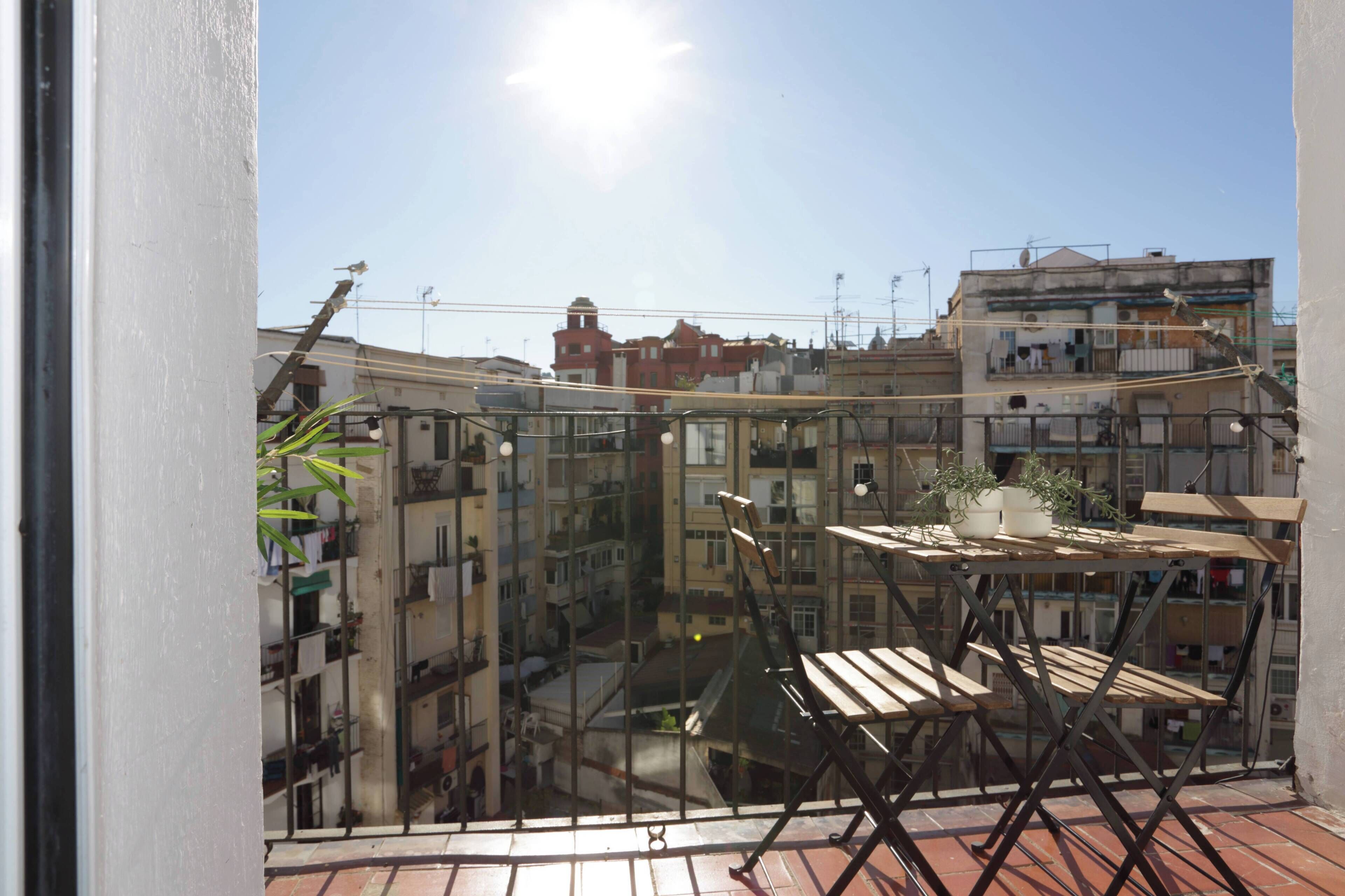 A balcony with a wooden table and chairs, a potted plant, and a view of a densely packed urban area with buildings.
