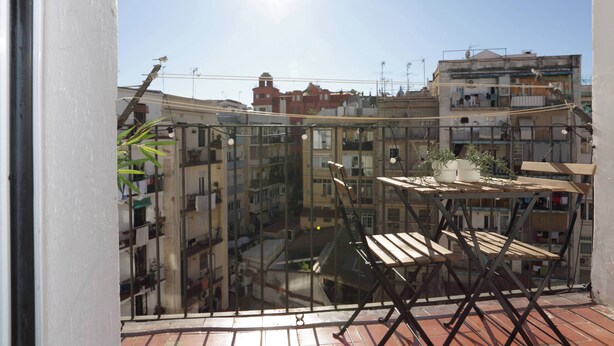 A balcony with a wooden table and chairs, a potted plant, and a view of a densely packed urban area with buildings.