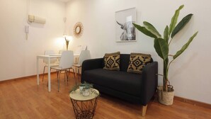 A living room with a black sofa, a dining table with chairs, a potted plant, and a wall-mounted light fixture.