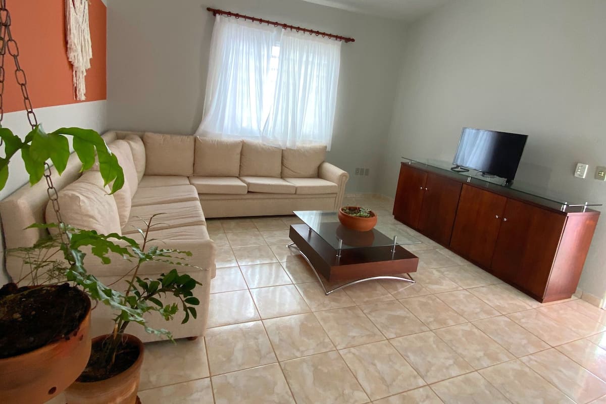 Ein Wohnzimmer mit einem beigefarbenen Sofa, einem Glastisch, einem Fernseher auf einer Holzkommode und einem Deckenventilator.