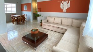 Un soggiorno con un divano beige, un tavolino da caffè in vetro con una pianta, un'area pranzo con sedie di legno e una ventola a soffitto.