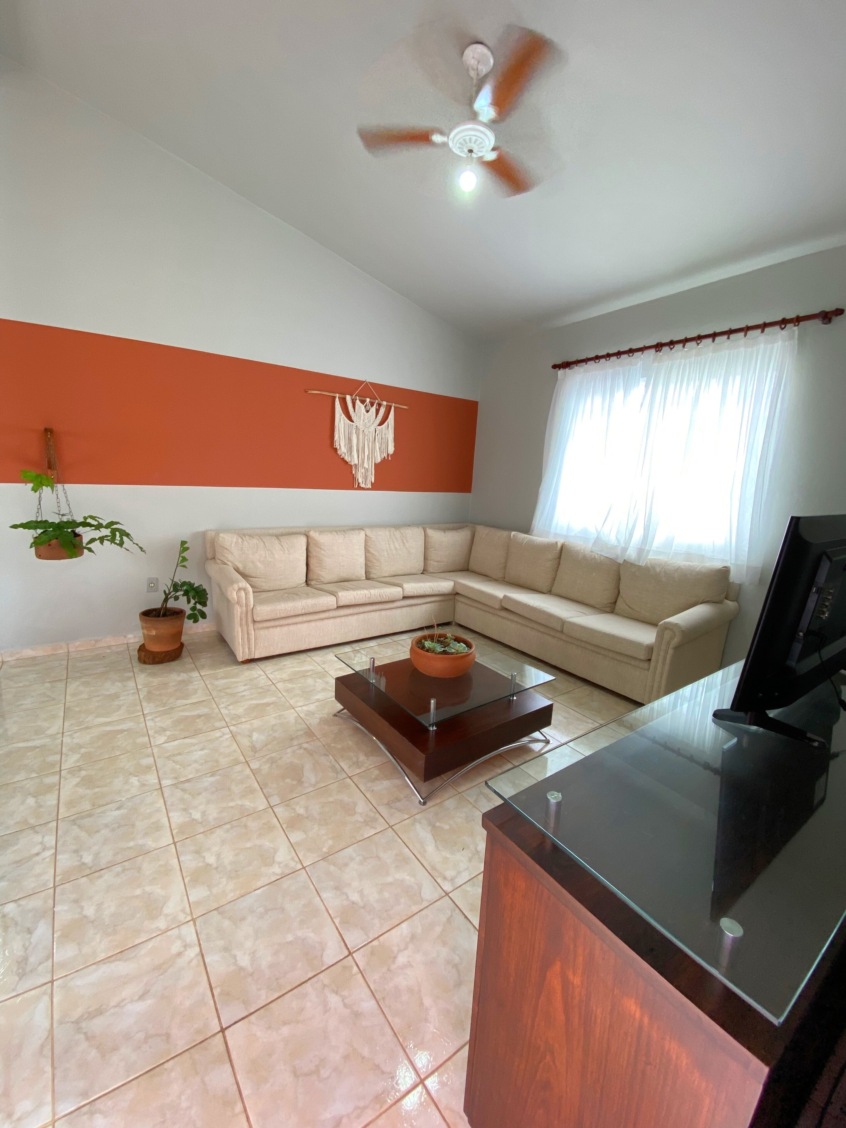 A living room with a beige sectional sofa, a glass coffee table, a ceiling fan, and a TV on a wooden stand.