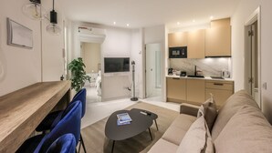Un salon moderne avec une table à manger en bois, des chaises bleues, une table basse noire et un canapé beige. On aperçoit, au fond, une cuisine avec des meubles beiges et un plan de travail en marbre.