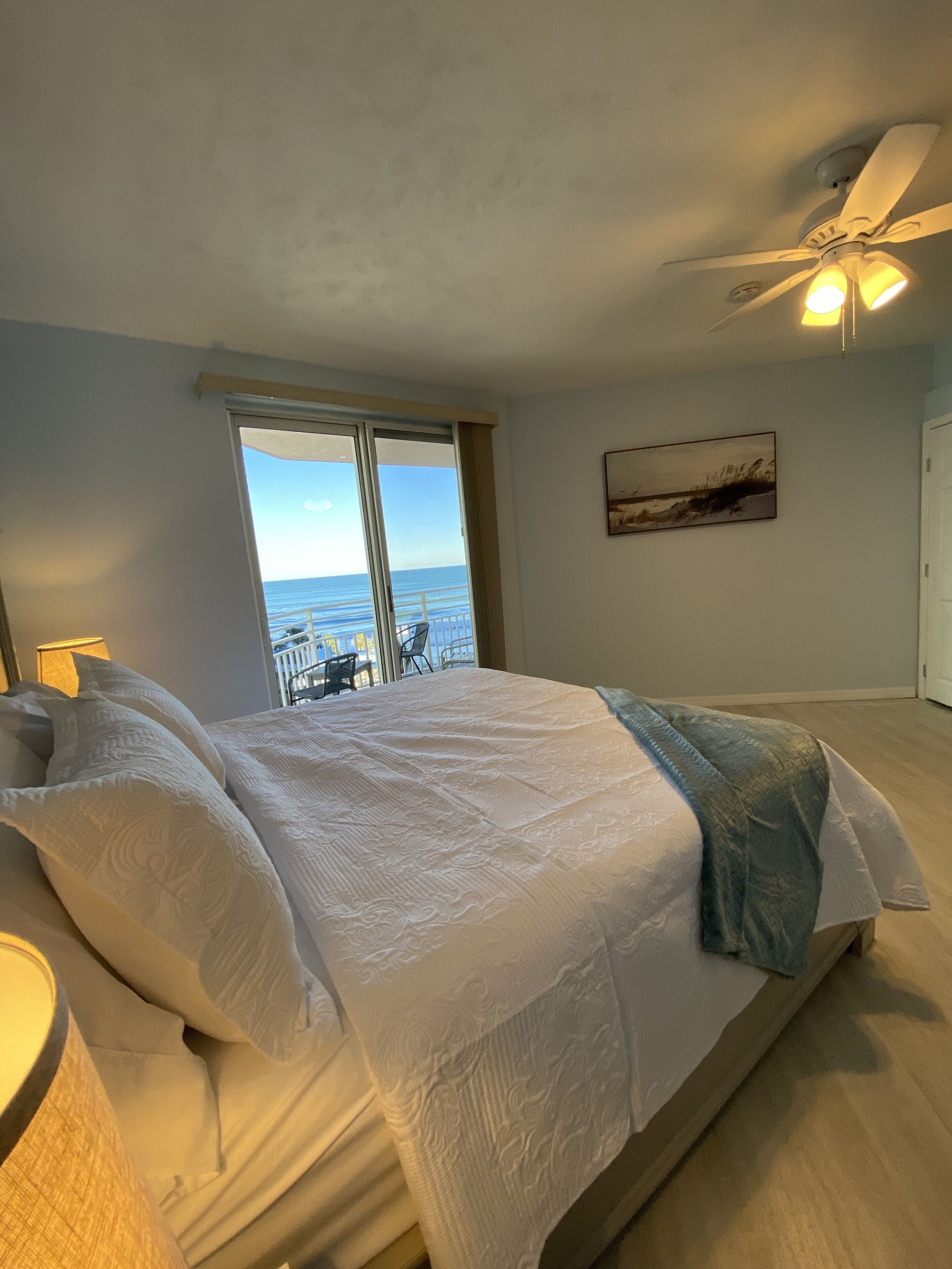 Un dormitorio con una cama grande, un ventilador de techo, una ventana con vista al mar y un cuadro en la pared.