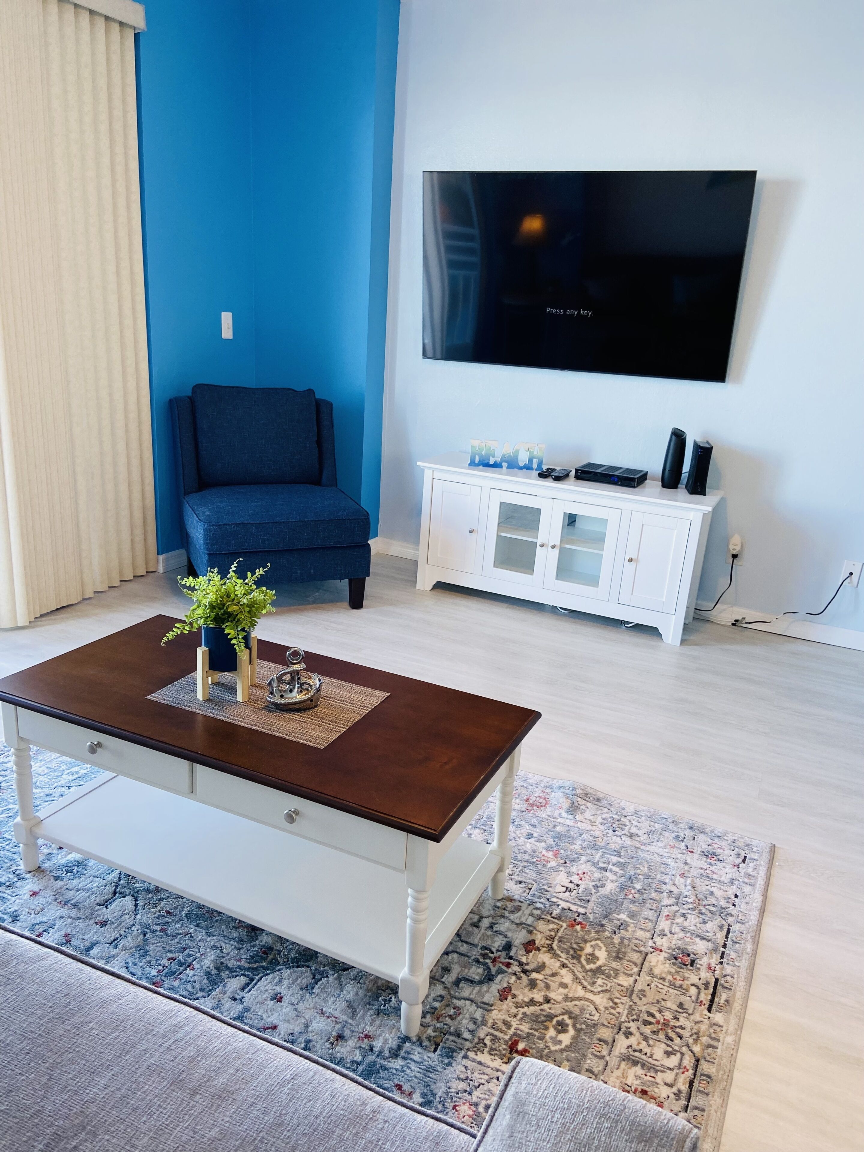 Een woonkamer met een blauwe wand, een grote flatscreen televisie bevestigd boven een witte meubelunit, een blauwe bank en een bijzettafeltje met decoratieve objecten.