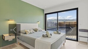 Ein modernes Schlafzimmer mit einem großen Bett, einem Nachttisch, einem Sessel und Blick auf einen Pool sowie Berge durch eine Glastür.