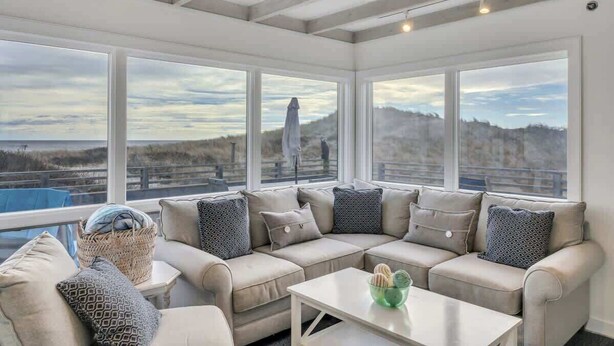 A sunroom with a large sectional sofa, a small table, and a view of the beach and hills. - Unit 511: Cottage | Ocean View | Nautilus (Amagansett)