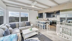 A modern living room with a sofa, coffee table, and dining area. A kitchen is visible with cabinets and appliances. Large windows offer a view of the outdoors. - Unit 511: Cottage | Ocean View | Nautilus (Amagansett)