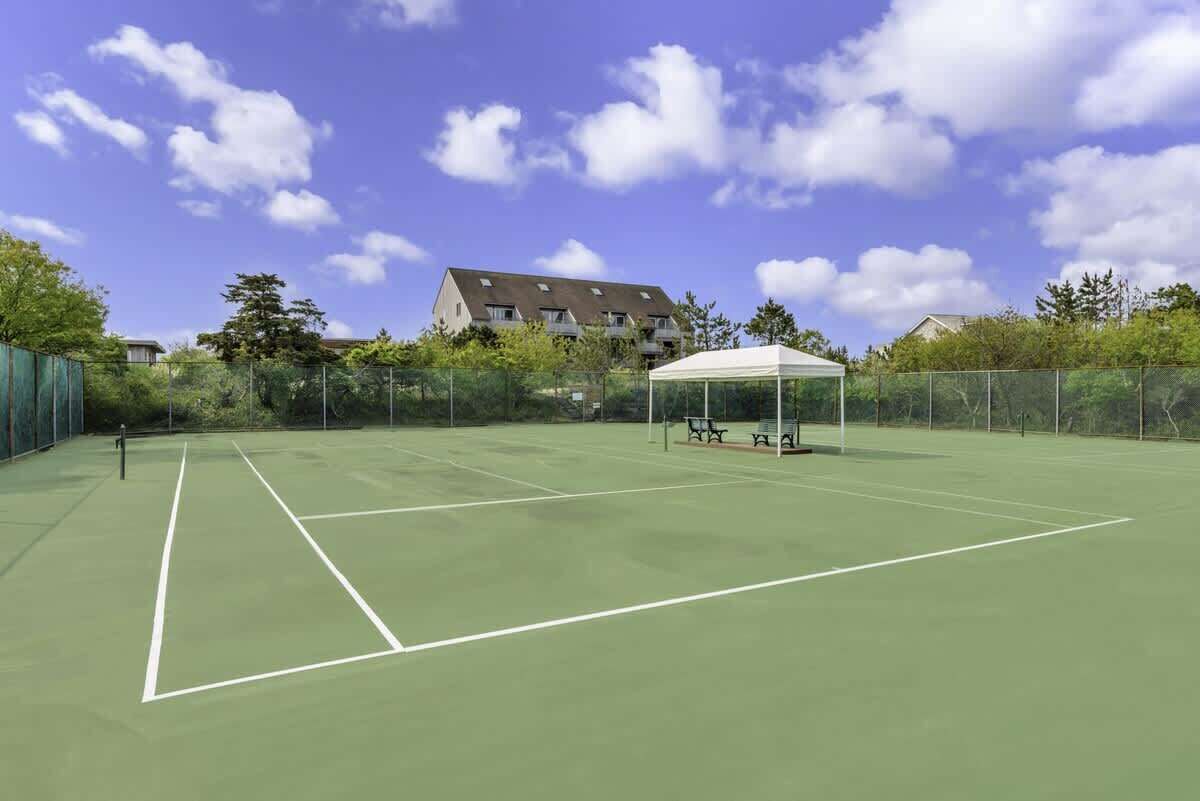 An outdoor tennis court with a white pavilion, surrounded by a fence and trees, with buildings in the background.