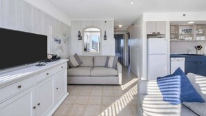 A modern living room with a flat-screen TV, a sofa, and a dining area with a kitchen in the background. - Unit 611: Two-Bedroom | 1st Floor-Dune View | Periwinkle (Amagansett)