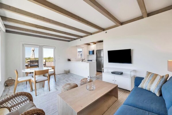 Una sala de estar moderna con un sofá azul, una mesa de centro de madera y sillas de mimbre. Se ve una cocina con televisión y armarios al fondo.