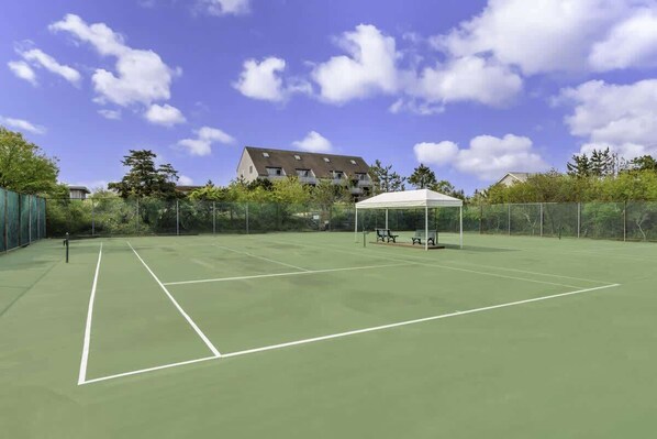 An outdoor tennis court with a white pavilion, surrounded by a fence and trees, with buildings in the background.