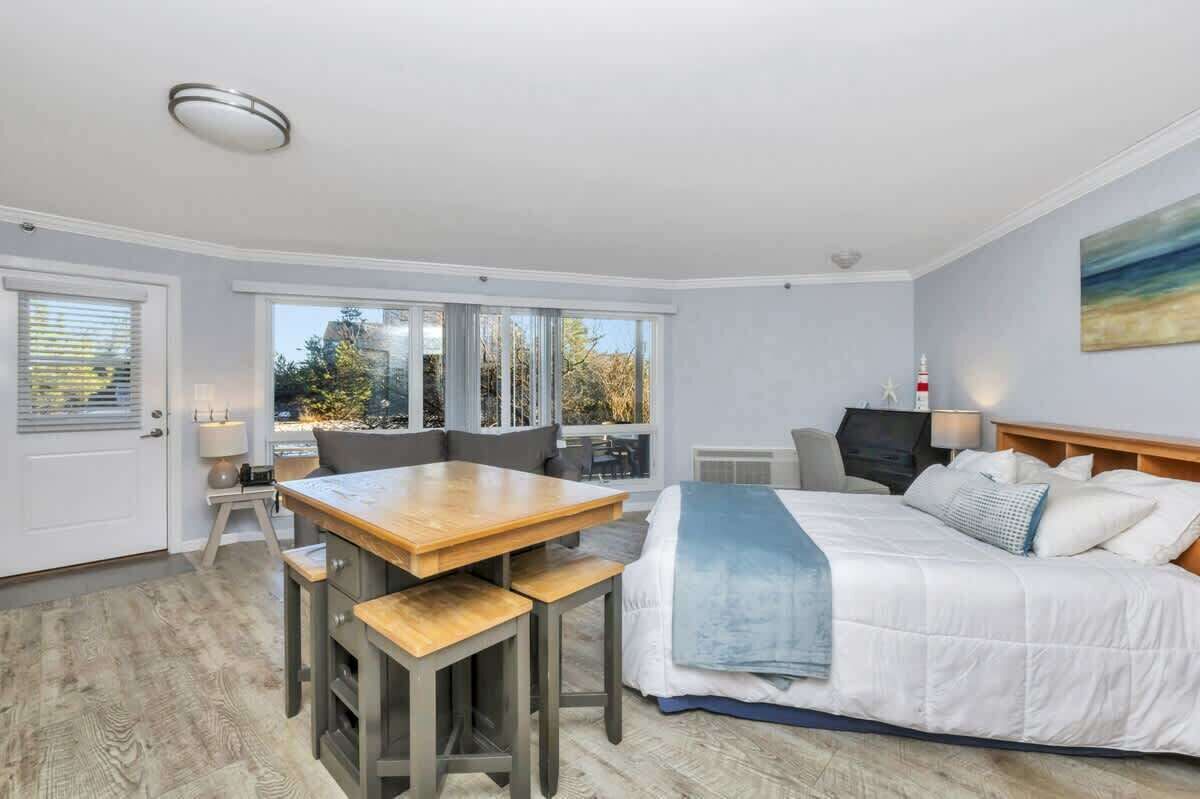 Una habitación de hotel moderna con una cama grande, una mesa de comedor con sillas, una pequeña cocina y amplias ventanas que ofrecen vistas al exterior.