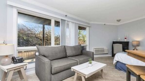 A modern hotel room with a grey sofa, a small white table, a bedside table with a lamp, and a view of trees through large windows.