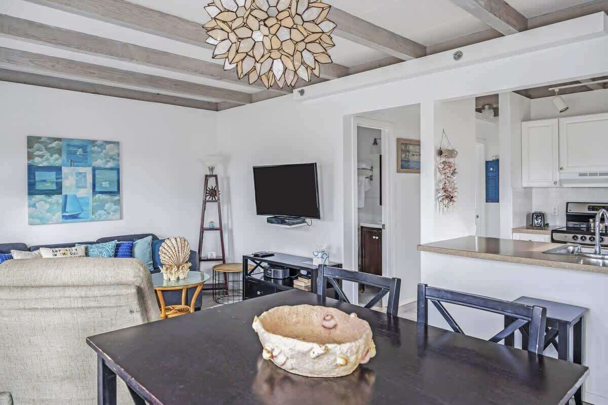 Una sala de estar moderna con una mesa de comedor de madera oscura, televisor y una cocina con encimera y lavamanos.