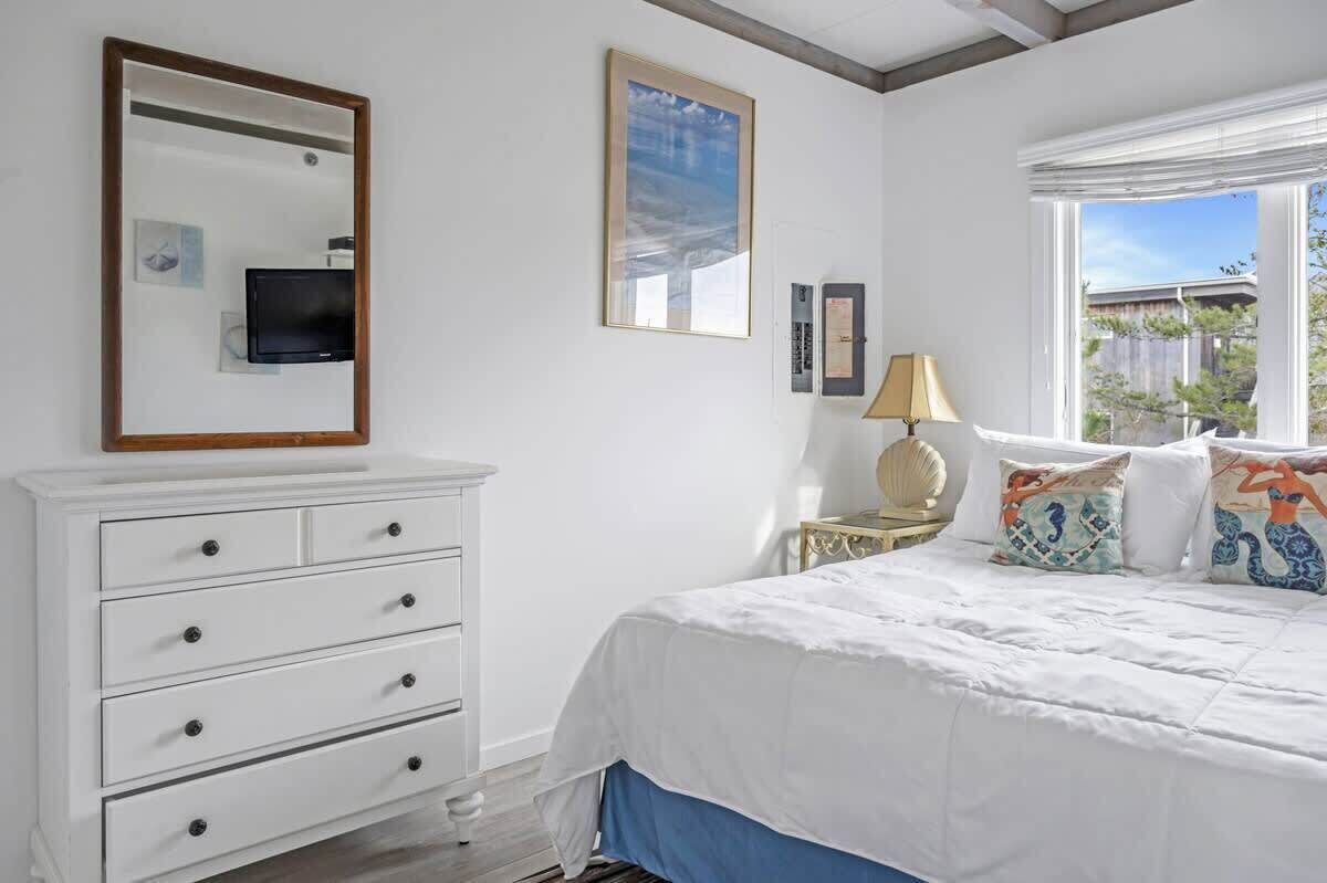 Un dormitorio con cama, tocador blanco, televisor, lámpara y ventana con vistas.