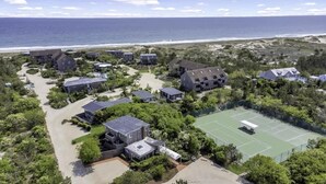 An aerial view of a coastal residential area with multiple houses, a tennis court, and a beach in the background.