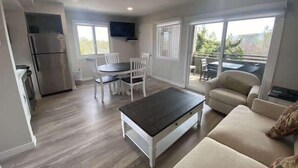 A modern living room with a sofa, a coffee table, a dining area with chairs, a kitchen with a refrigerator, and large windows.