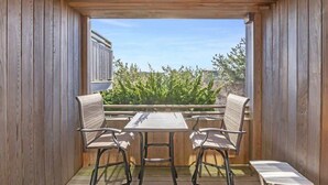 Un terrazzo in legno con un tavolo e due sedie, affacciato su un giardino con arbusti verdi e un cielo sereno.