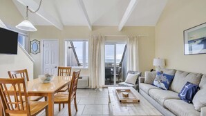 Amplia sala de estar con una mesa de comedor de madera, sillas, sofá y una mesa de centro. Hay un televisor montado en la pared, una ventana con cortinas y una puerta que da a otra habitación.