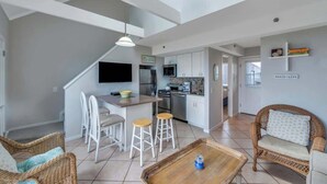 A modern kitchen with white cabinets, a dining area with white chairs, and a wicker sofa. - Unit 1722: 1-Bedroom with Loft | 2nd Floor-Ocean Front Ocean View | Ocean Edge (Amagansett)
