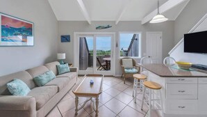 A modern living room with a sofa, coffee table, and a dining area with chairs. There is a large window providing a view of the outdoors. - Unit 1722: 1-Bedroom with Loft | 2nd Floor-Ocean Front Ocean View | Ocean Edge (Amagansett)