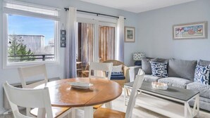 Una sala de estar con una mesa de comedor, sillas, un sofá y una mesa de centro. Se observa una ventana con cortinas, una puerta y vistas al exterior.