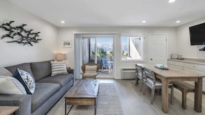 A modern living room with a sofa, coffee table, and dining area. There is a view through the glass door to the outside. - Unit 1816: 1-Bedroom | 1st Floor-Garden View | Dunes East (Amagansett)