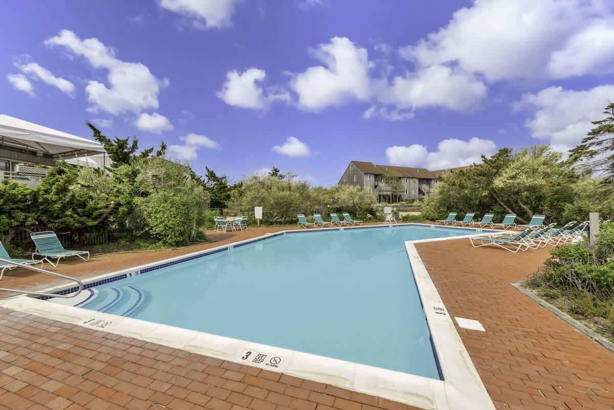 Une piscine rectangulaire dont le bord est recouvert de carreaux, entourée d’une terrasse en briques. Des chaises longues sont disposées le long du bord de la piscine, et on aperçoit un bâtiment avec un toit en pente à l’arrière-plan.