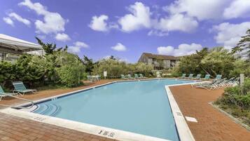 Une piscine rectangulaire dont le bord est recouvert de carreaux, entourée d’une terrasse en briques. Des chaises longues sont disposées le long du bord de la piscine, et on aperçoit un bâtiment avec un toit en pente à l’arrière-plan.