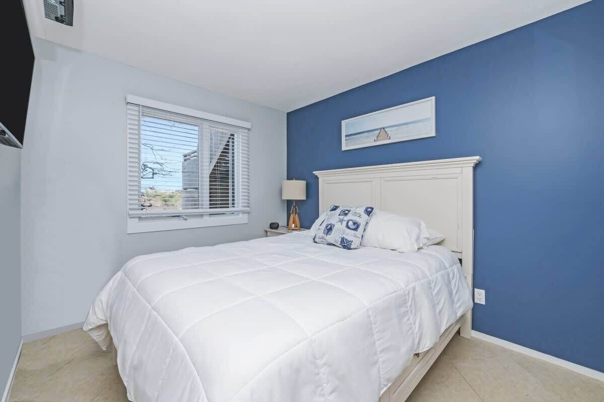 Quarto com cama, janela com persianas, mesa de cabeceira com abajur e um quadro na parede.