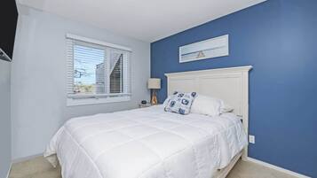 Quarto com cama, janela com persianas, mesa de cabeceira com abajur e um quadro na parede.