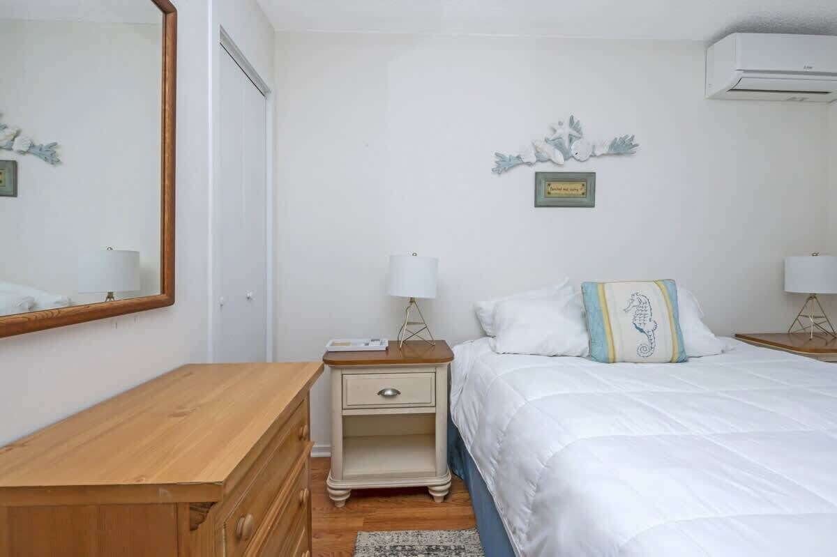 A bedroom with a wooden dresser, a nightstand with a lamp, a bed with white bedding and a decorative pillow, a wall-mounted air conditioner, and a framed picture.
