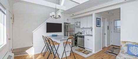 A modern apartment with a kitchen, dining area, and living room. It features a loft-style ceiling, a flat-screen TV, and a kitchenette.