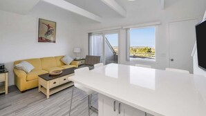 A modern living room with a yellow sofa, a white dining table, and a large window offering a view of the city. - Unit 2022: 1-Bedroom with Loft | 2nd Floor-Bay View | Court Side (Amagansett)