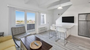 A modern living room with a sofa, a coffee table with a remote, a TV, a kitchen area with a fridge, and large windows with a view. - Unit 2022: 1-Bedroom with Loft | 2nd Floor-Bay View | Court Side (Amagansett)