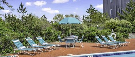 Un espace piscine avec des chaises longues et une table placée sous un parasol, entouré de verdure et avec un bâtiment en arrière-plan.